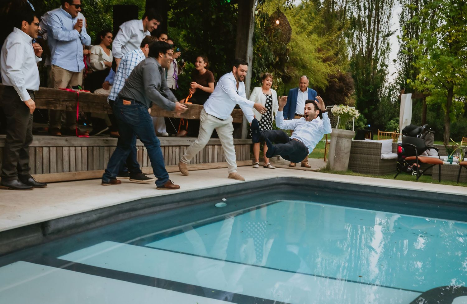 Men playfully pushing a guest into a swimming pool at an outdoor party with onlookers laughing nearby.