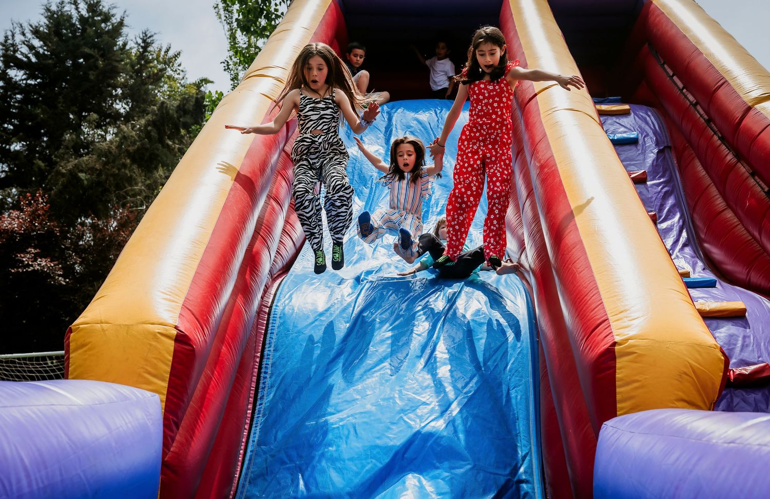 Three girls joyfully sliding down a colorful inflatable bounce house slide at an outdoor party.