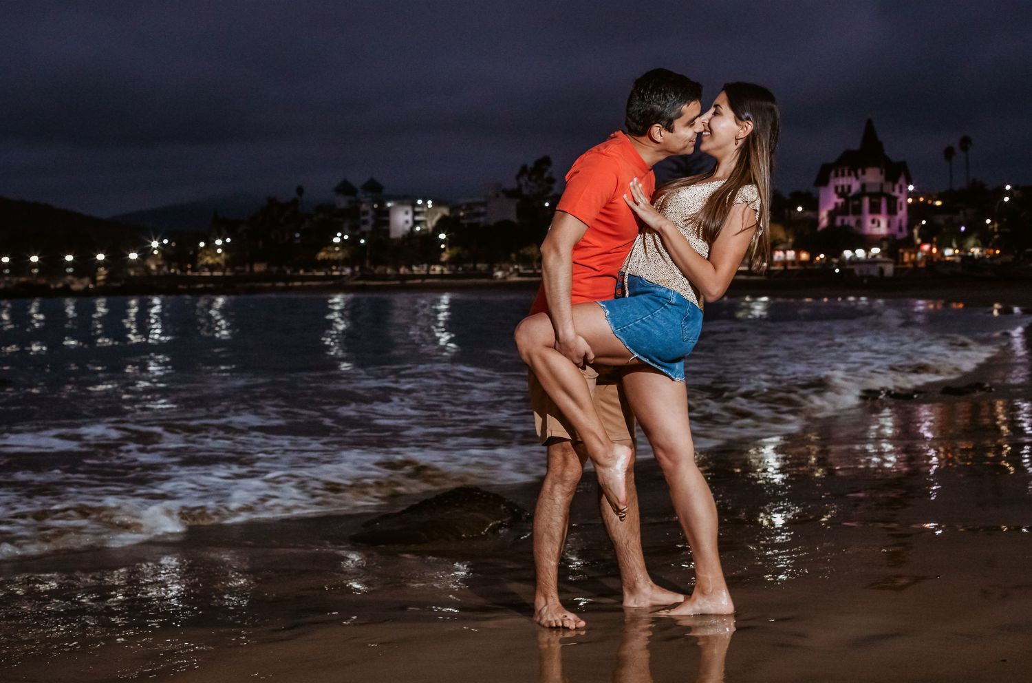 Couple sharing a kiss on a wet beach at night with glowing city lights behind them.