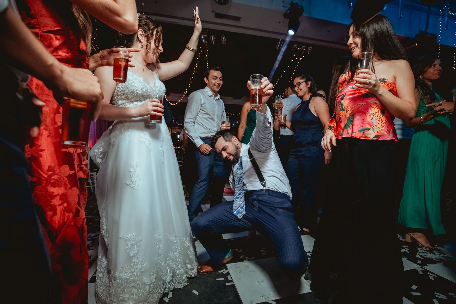 Energetic wedding reception dance floor scene with bride, groom, and guests celebrating with drinks.