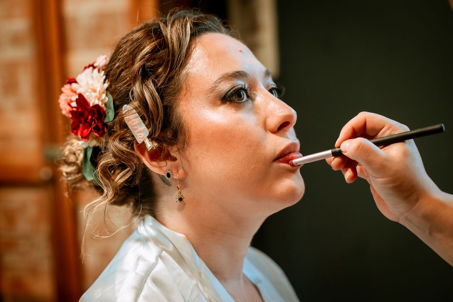 Makeup artist applying lipstick to bride with floral hair accessories during wedding preparations.