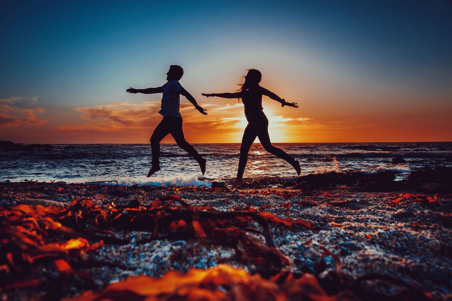 Two women jumping joyfully on a rocky beach at sunset with vibrant orange and blue sky.