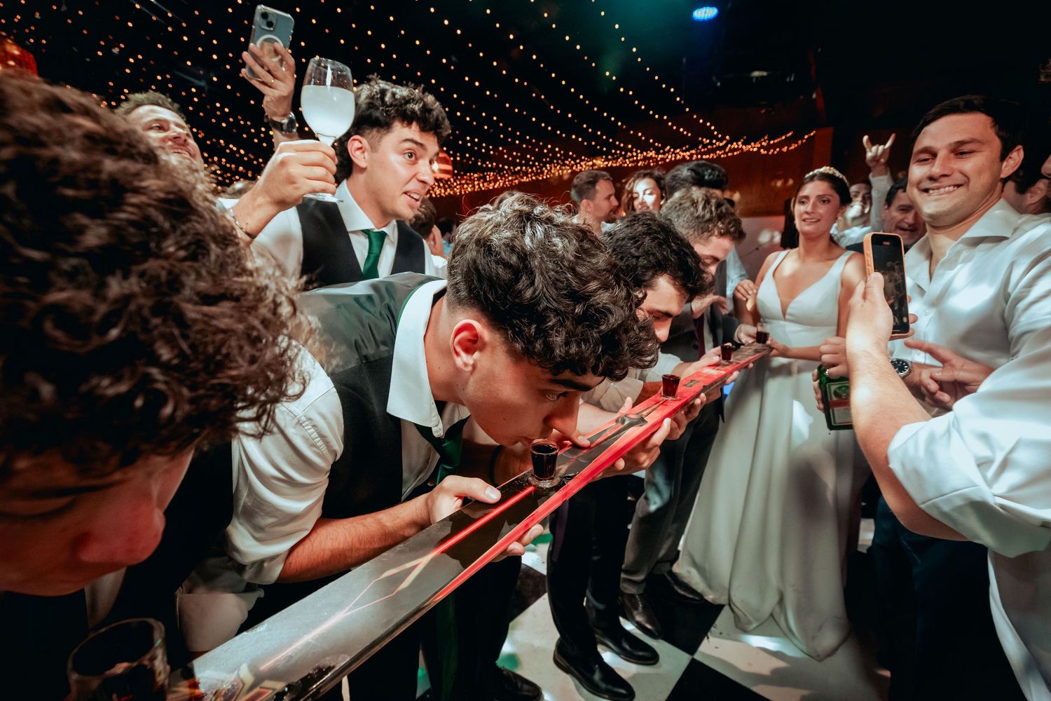 Groom and guests play drinking game at wedding reception, leaning over table with red shot tubes.