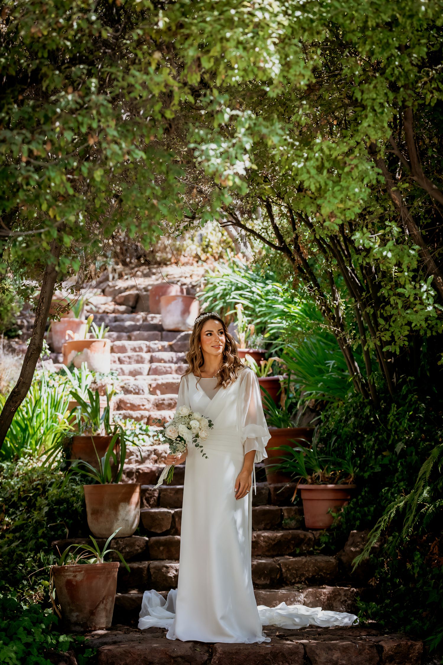Bride in a flowing white gown holds a bouquet while standing on stone steps in a green garden.