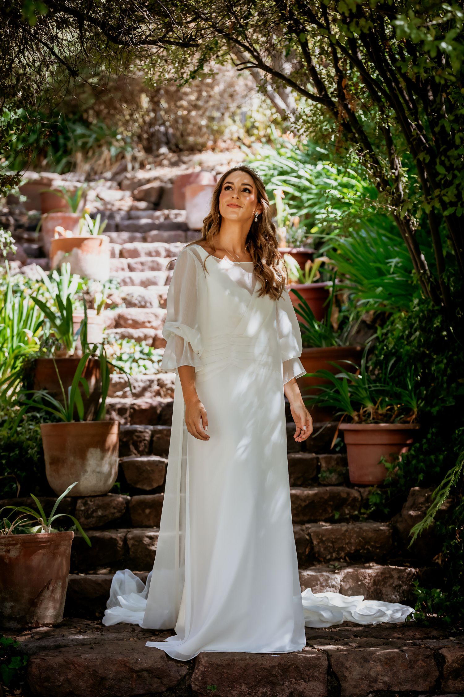 Beautiful bride in a white gown with sheer sleeves poses on stone steps in a green garden.