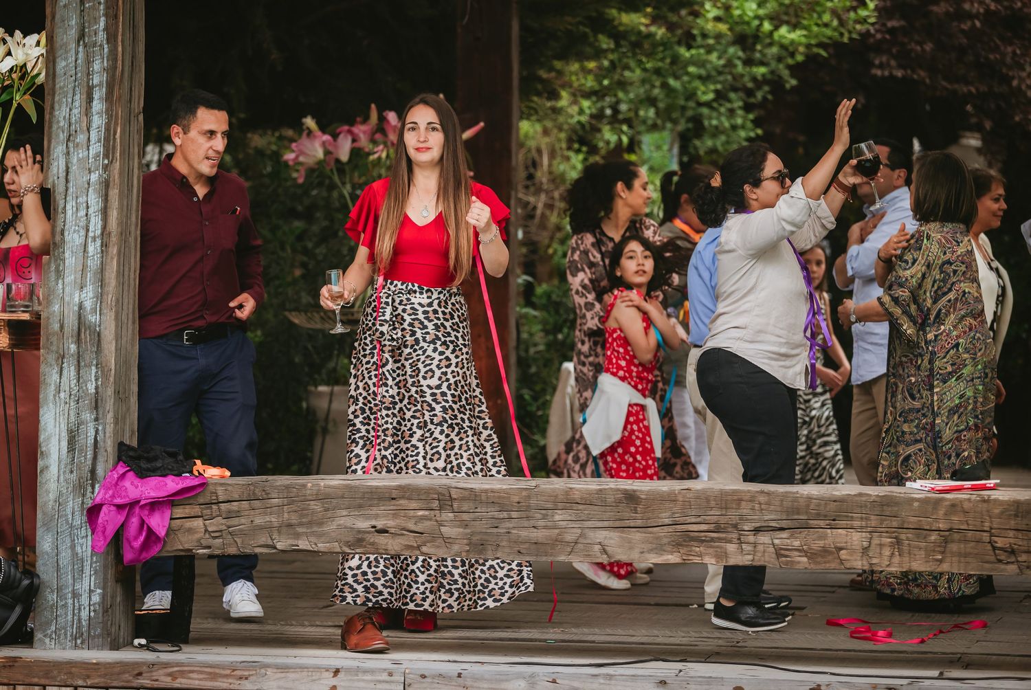 People in festive attire gather on a wooden porch at an outdoor celebration, with colorful decorations nearby.