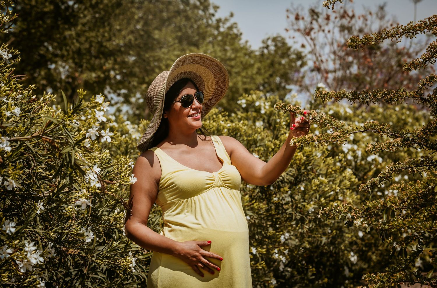 Pregnant woman in yellow dress and sun hat smiling among flowering trees outdoors.