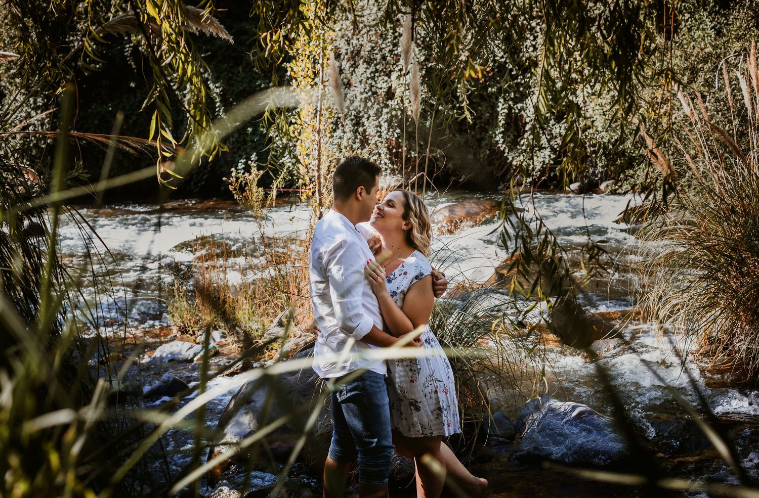 Couple embracing romantically beside a flowing creek surrounded by lush greenery and rocks in a natural outdoor setting.
