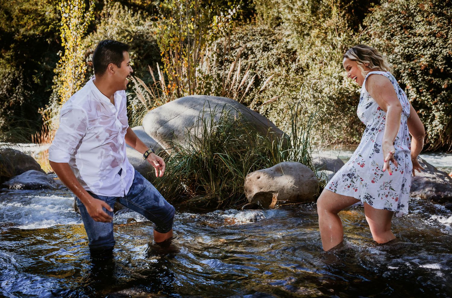 Couple wading in a rocky stream with a small animal nearby, surrounded by lush greenery.