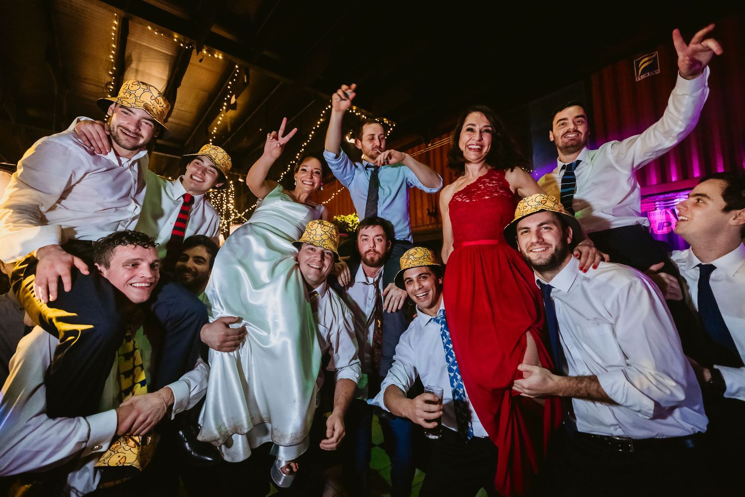 Wedding party group photo with bride in white, guests in crowns, celebrating joyfully at a festive reception.