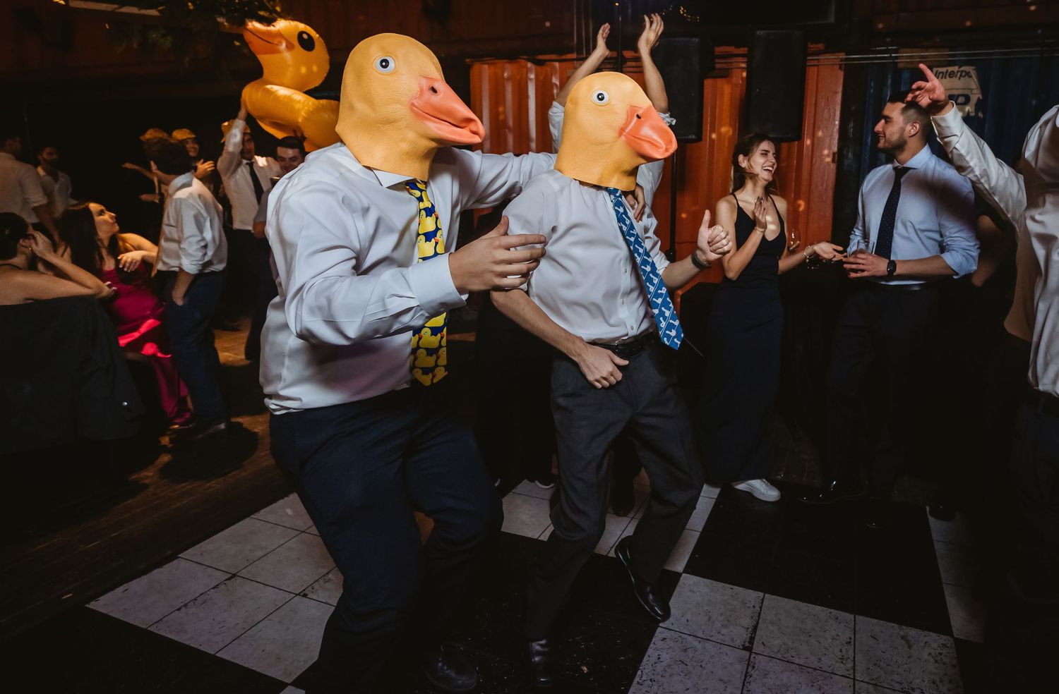 Two people wearing duck masks and suits dance energetically at a lively party event.