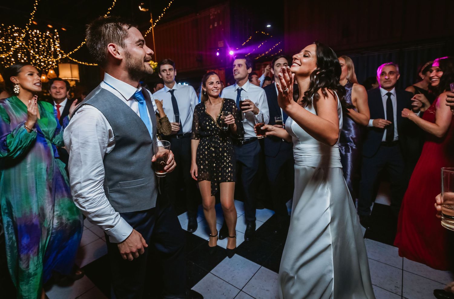 Bride and groom laughing and dancing together at their wedding reception surrounded by guests on a white dance floor.