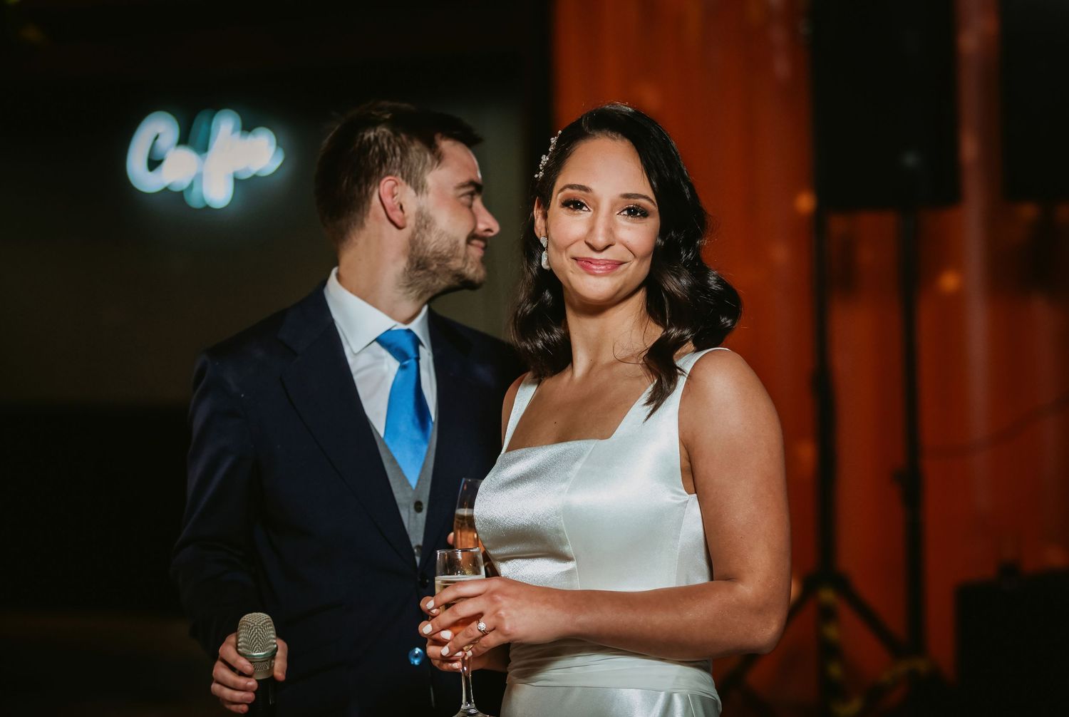 Bride and groom smiling together at wedding reception, holding champagne glasses, with blue neon sign in background.