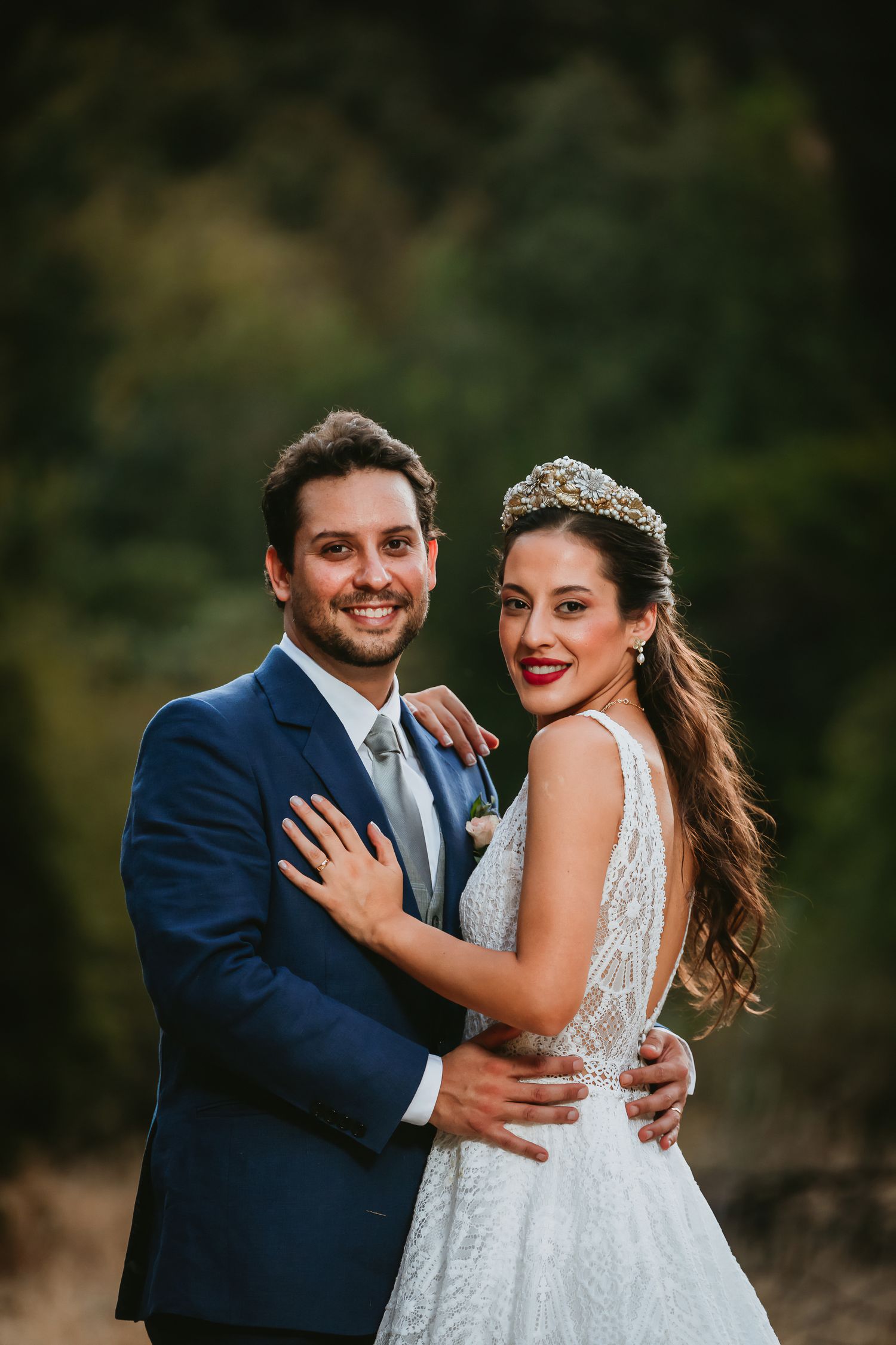 A smiling bride in a white gown and tiara embraces a groom in a navy suit outdoors in a romantic wedding portrait.