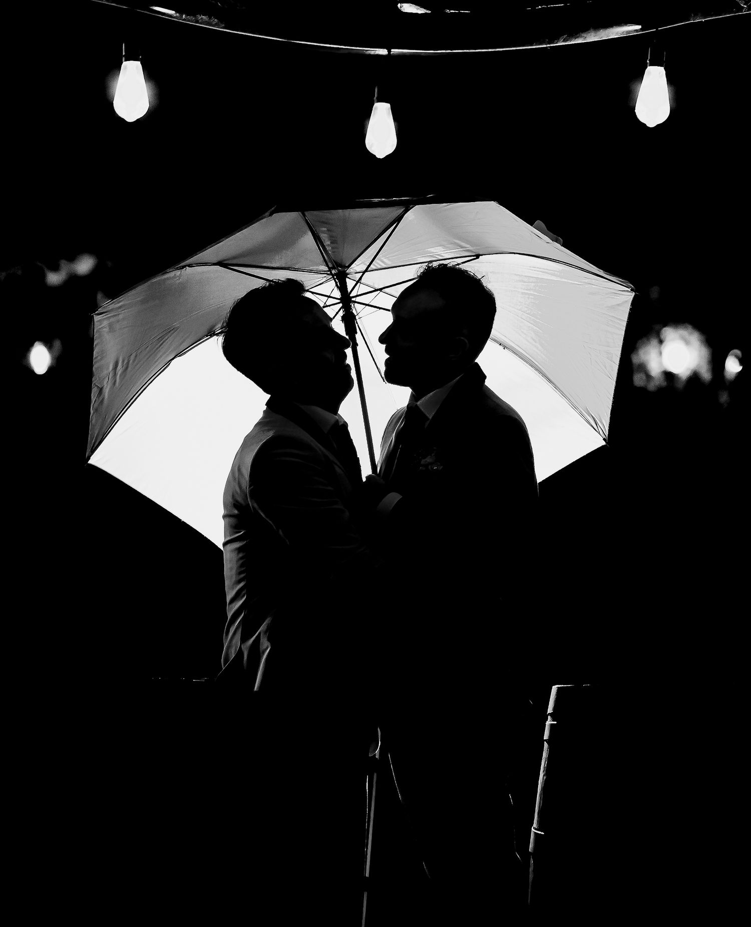 Black and white silhouette of two grooms sharing a romantic moment under a glowing umbrella at their wedding.