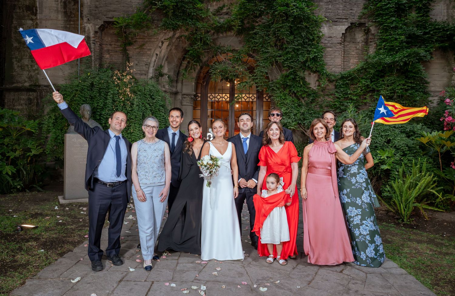 Wedding party poses with Chilean and Catalan flags outside historic stone building after ceremony.