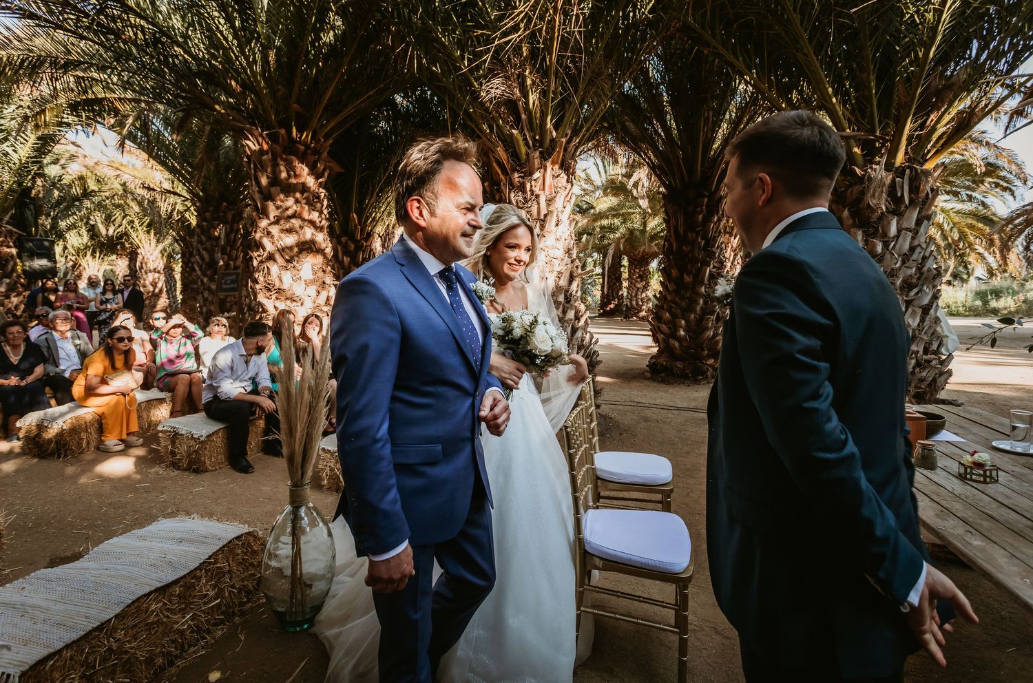 Father walking bride down palm-lined outdoor aisle toward waiting groom at altar.