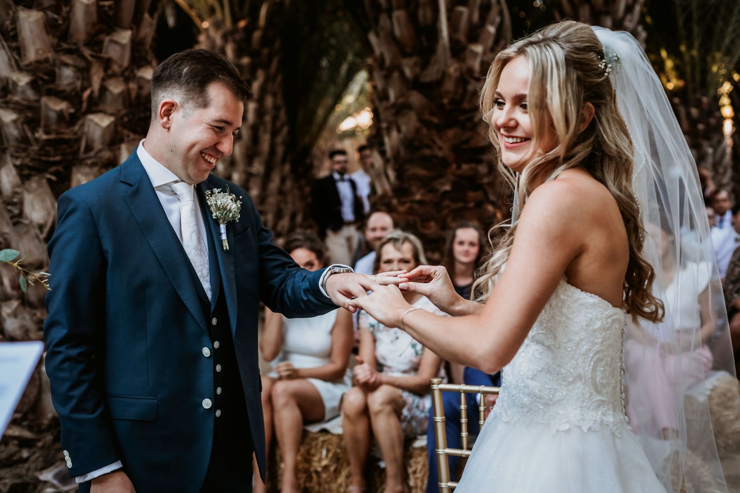 Smiling bride places a wedding ring on her grooms finger during their outdoor wedding ceremony.