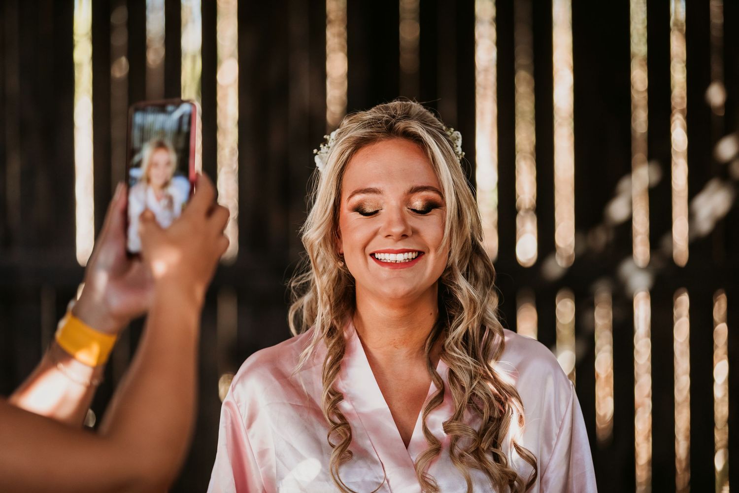 Smiling bride with floral hair accessories in pink robe being photographed on wedding day.