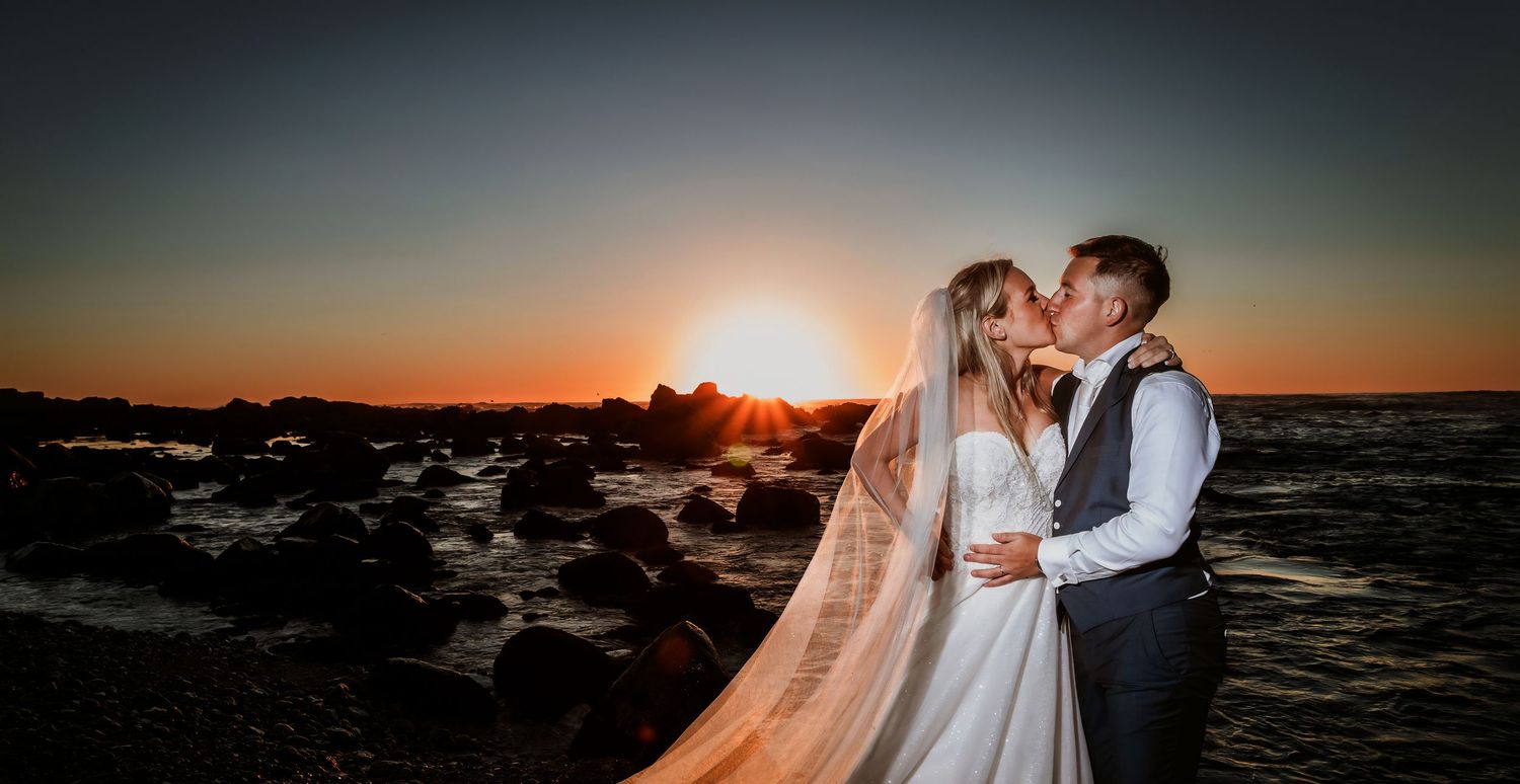 Bride and groom sharing a romantic kiss on rocky coast with vibrant sunset backdrop.