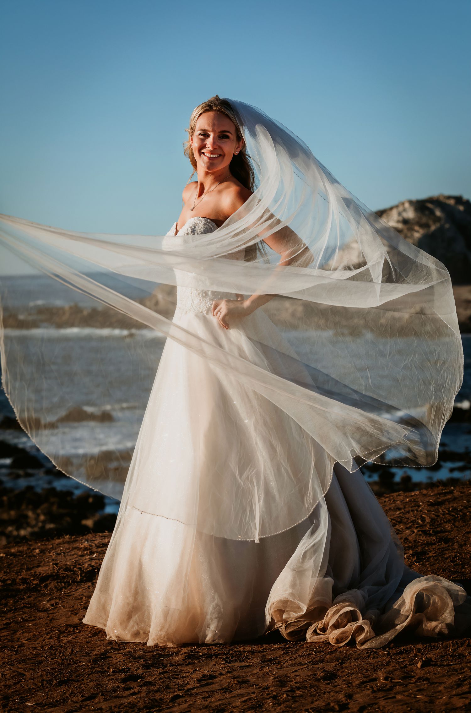 Smiling bride in strapless wedding gown with flowing veil caught in breeze on sunny rocky beach.