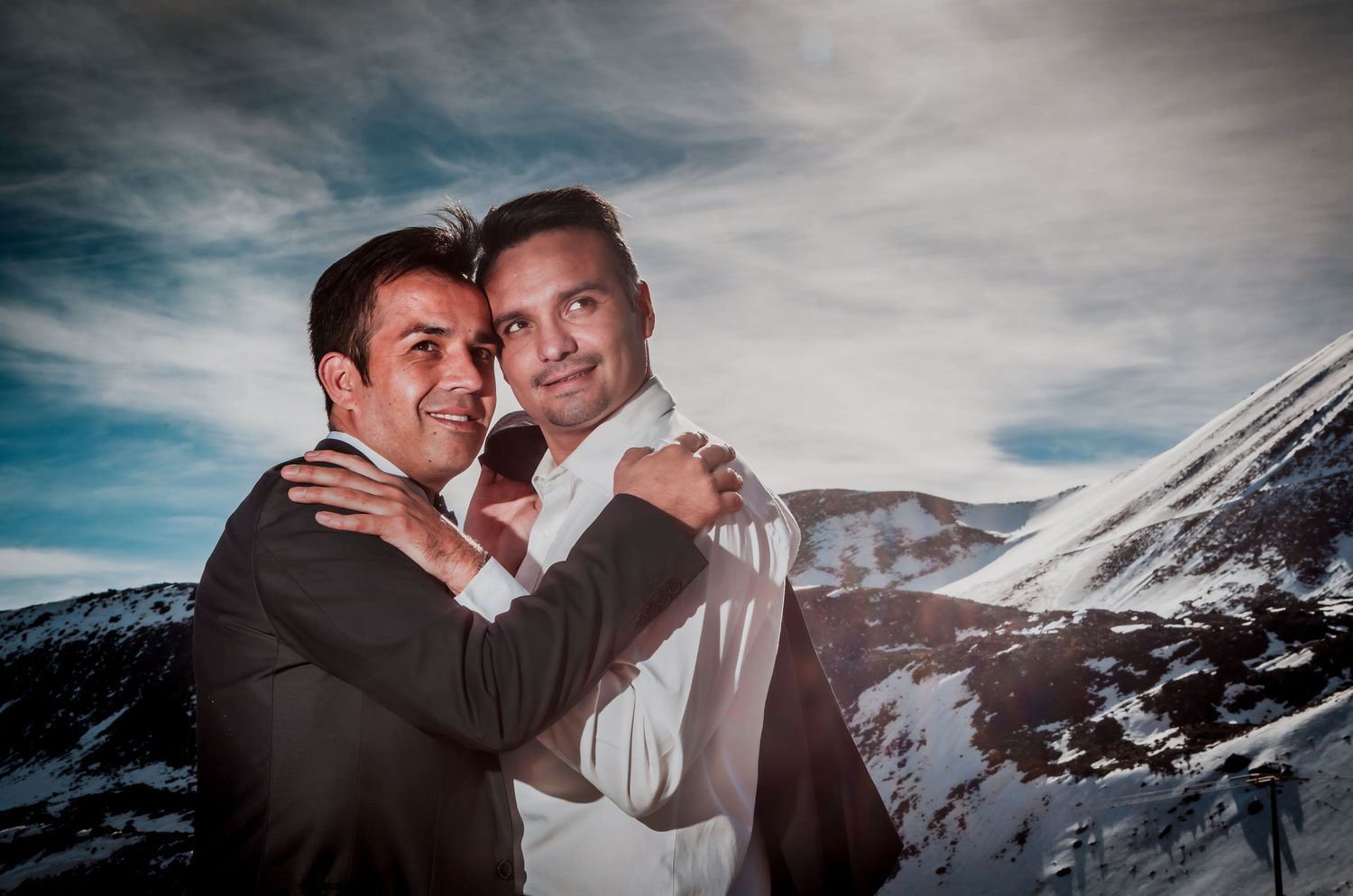 Two grooms embrace tenderly against a stunning snow-covered mountain landscape under a dramatic cloudy sky.