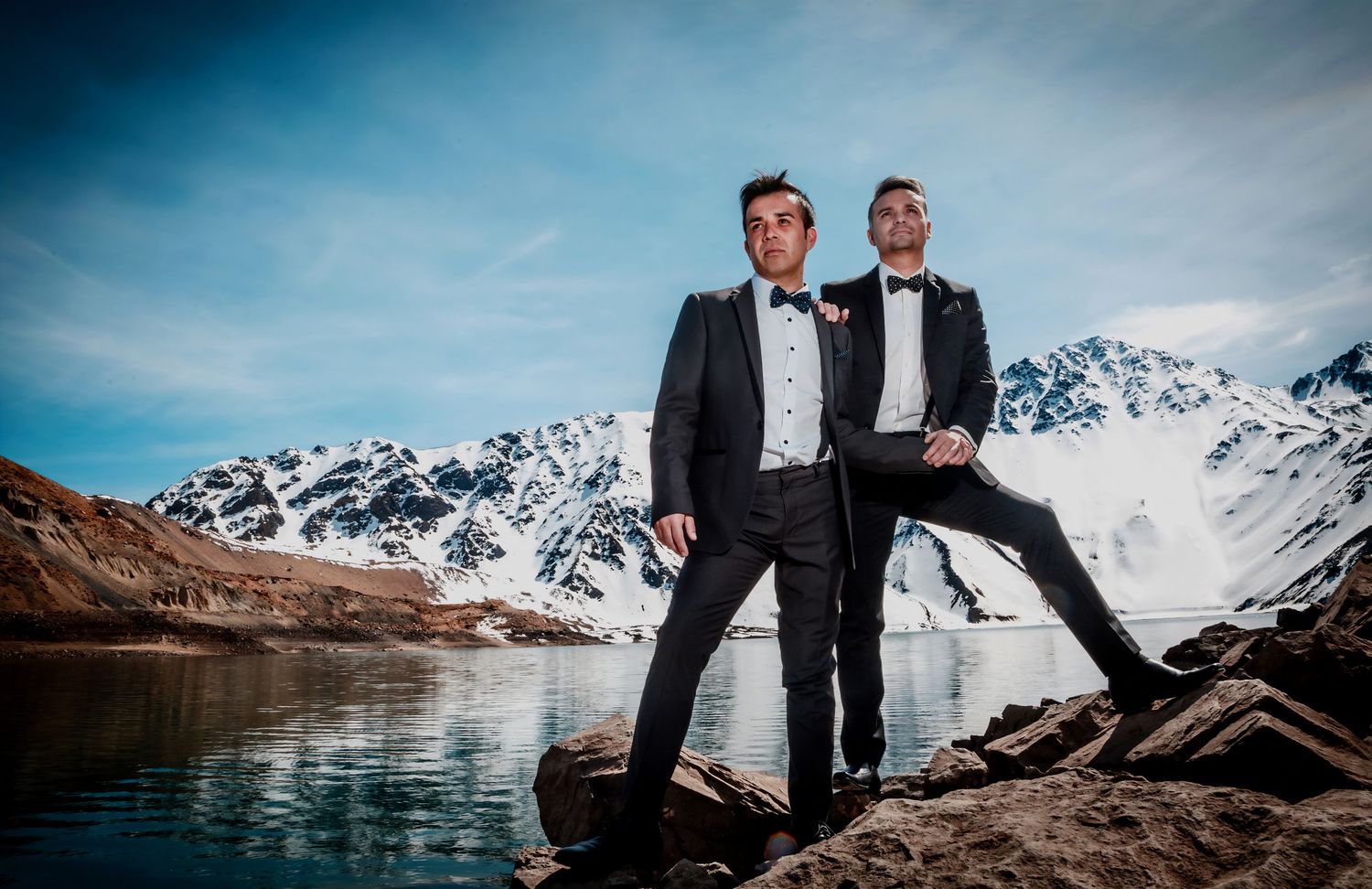Two grooms in elegant tuxedos pose on rocks with a breathtaking snowy mountain lake backdrop.