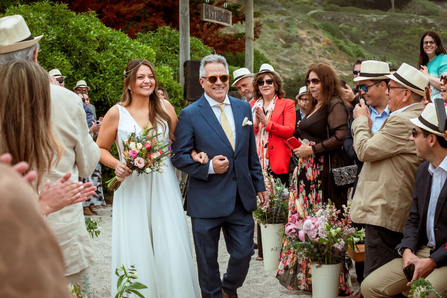 Bride walks down the aisle with her father as guests applaud at outdoor wedding.