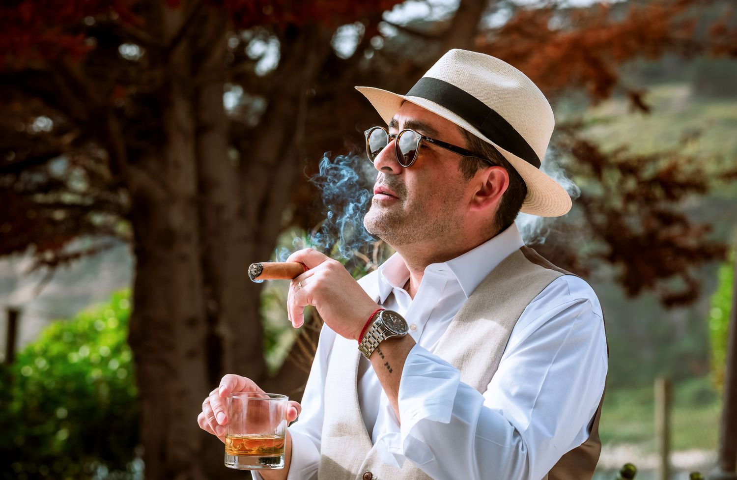 Stylish man in panama hat and sunglasses smoking a cigar while holding a glass of whiskey outdoors.