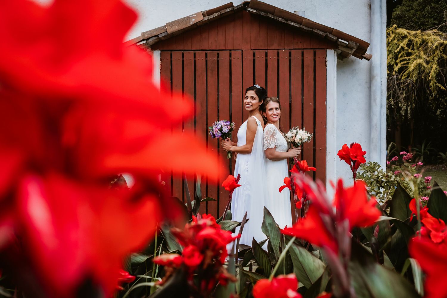 Two brides holding bouquets posing near red barn door surrounded by vibrant red tropical flowers.