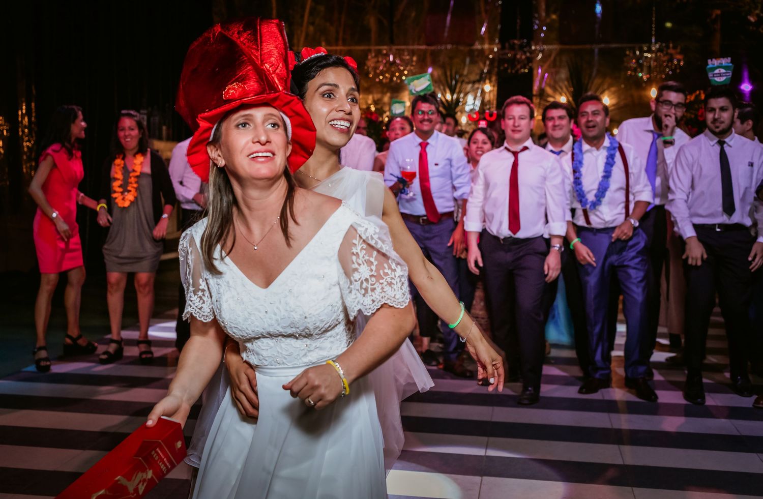 Two brides dance joyfully together, one in a large red hat, surrounded by wedding guests on the dance floor.