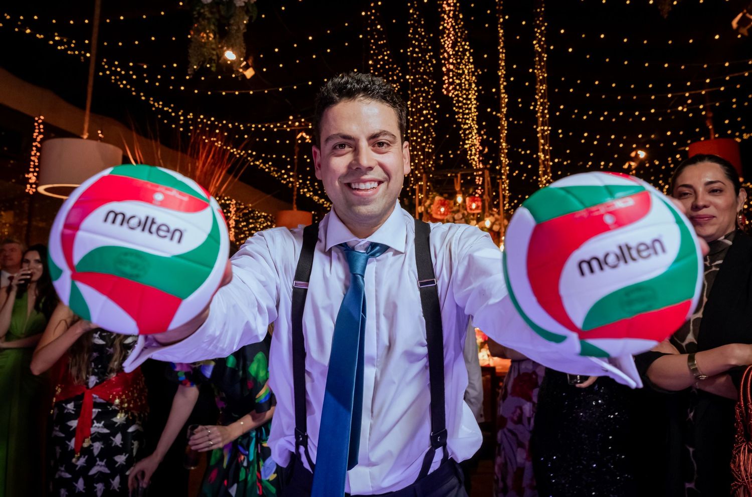 Smiling man holds two Molten volleyballs at outdoor wedding reception with string lights.
