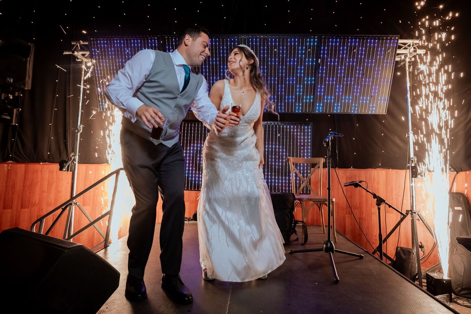 Bride and groom laugh together on stage surrounded by sparkling cold fireworks at wedding reception.