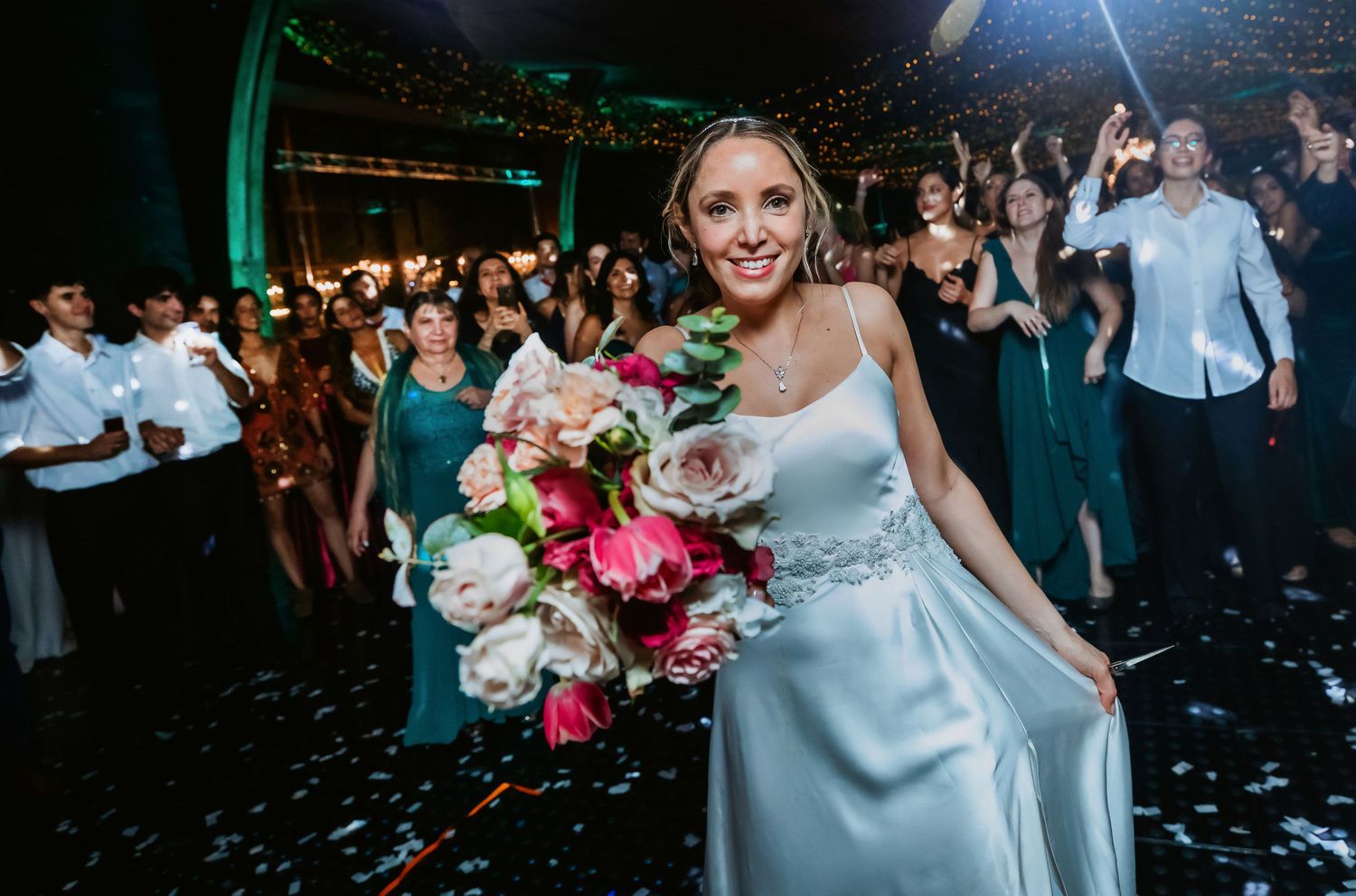 Smiling bride holds a pink and white bouquet on a confetti-filled wedding dance floor.