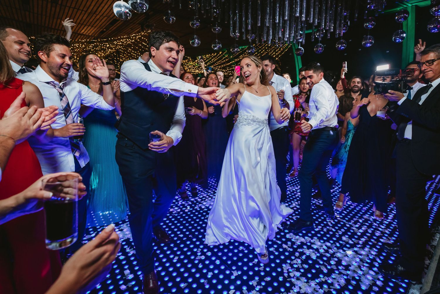 A bride and groom dance joyfully together on a glittering blue dance floor surrounded by applauding wedding guests.