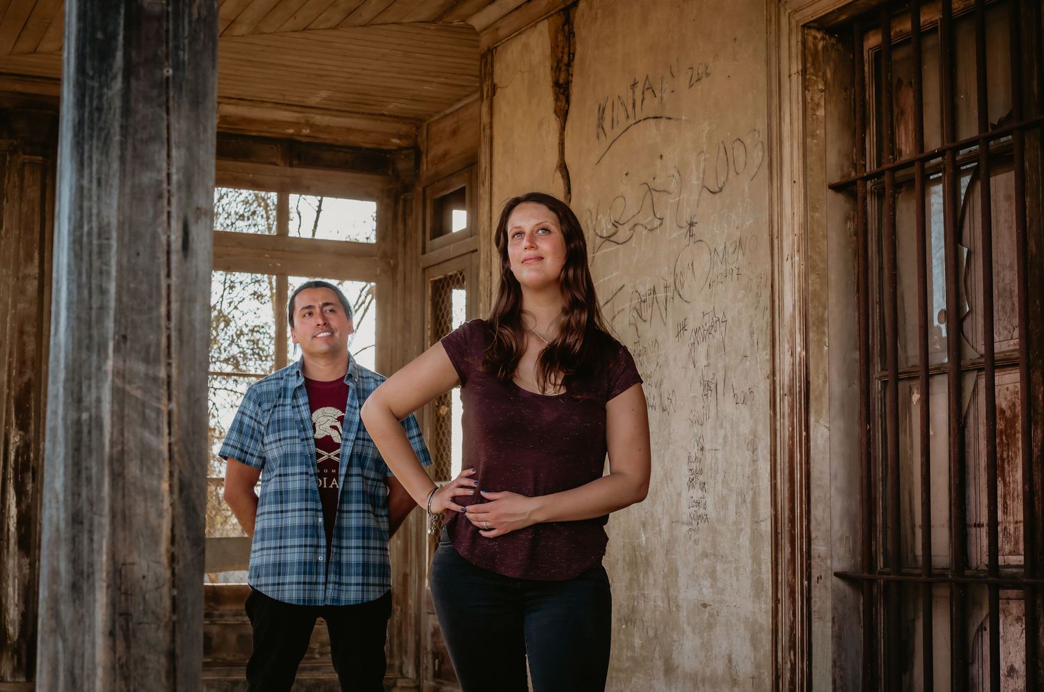 Couple standing in rustic abandoned building with weathered walls and large window behind them.