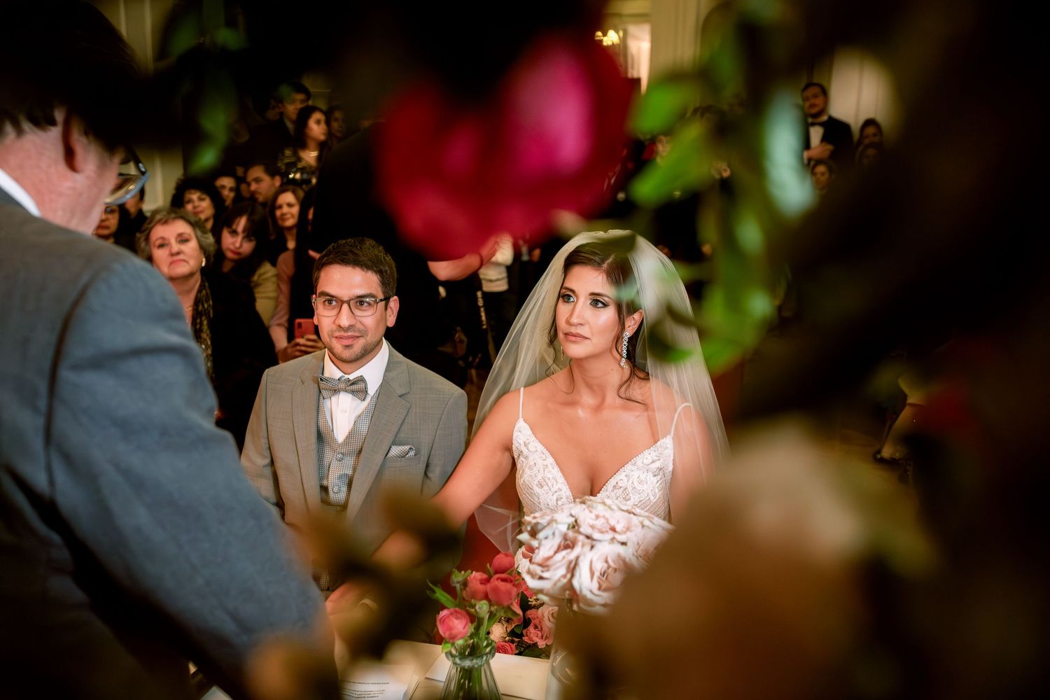 Bride and groom stand at wedding altar surrounded by roses and guests during elegant indoor ceremony.