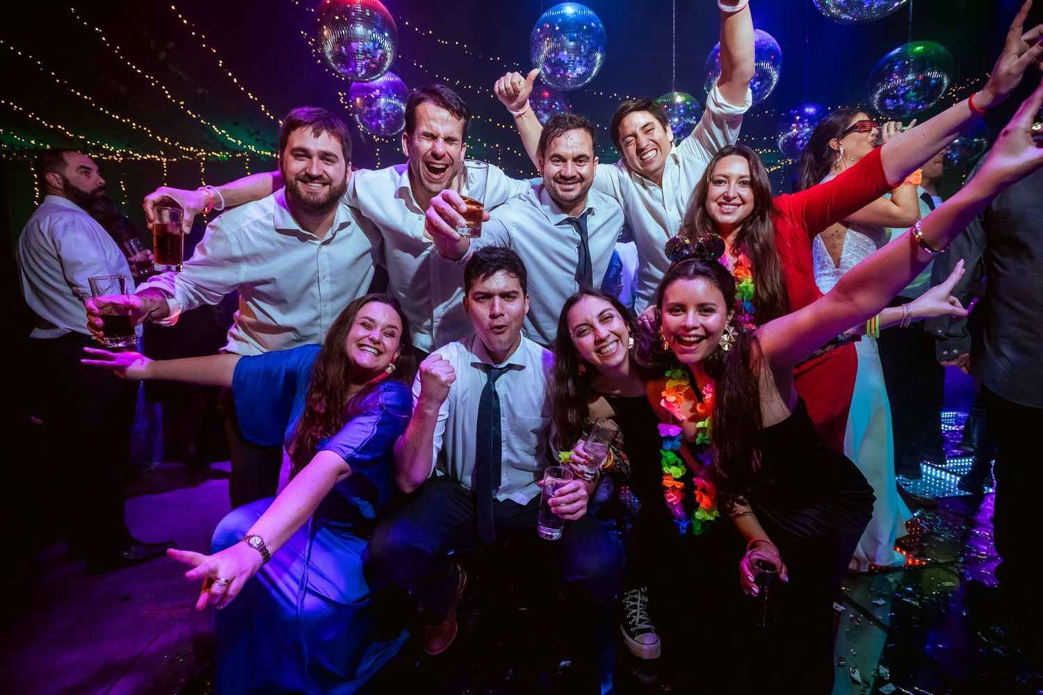 Group of young adults posing together at a colorful disco party with mirror balls and neon lights.