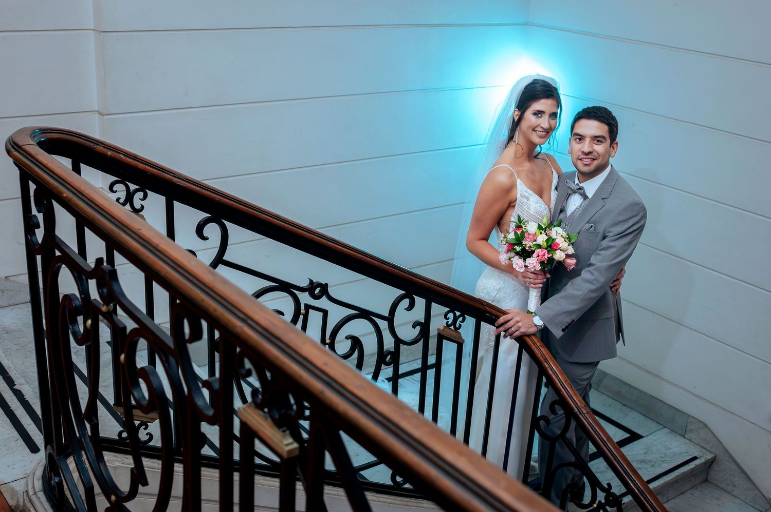 Bride and groom pose elegantly on ornate staircase, bride holding bouquet, groom in gray suit, blue light backdrop.