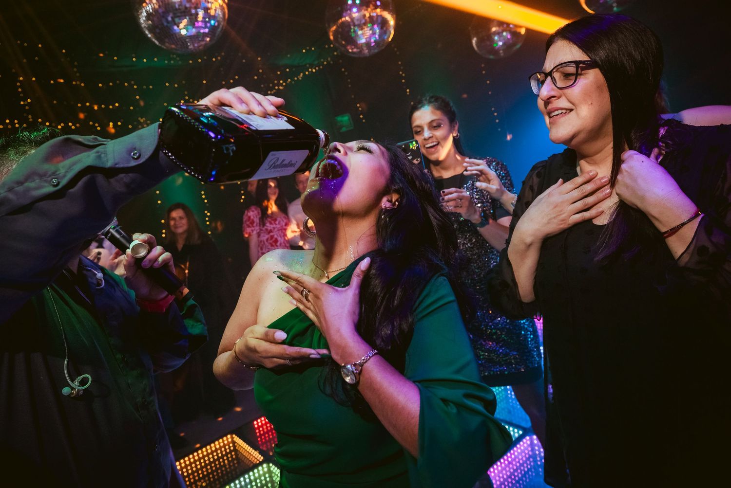 Women partying at a nightclub, one drinking from a bottle poured by another person under disco lights.