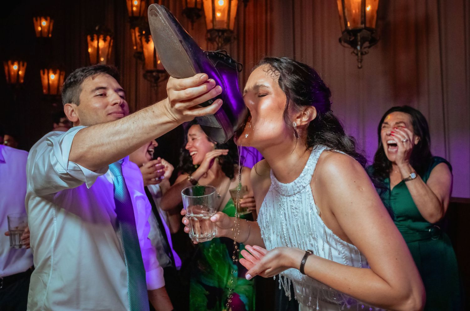Bride drinks from a large shoe held by a guest while onlookers react with surprise at reception.