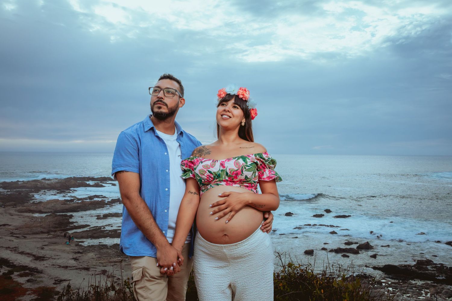 Couple holding hands by ocean, pregnant woman in floral crop top with flower crown.