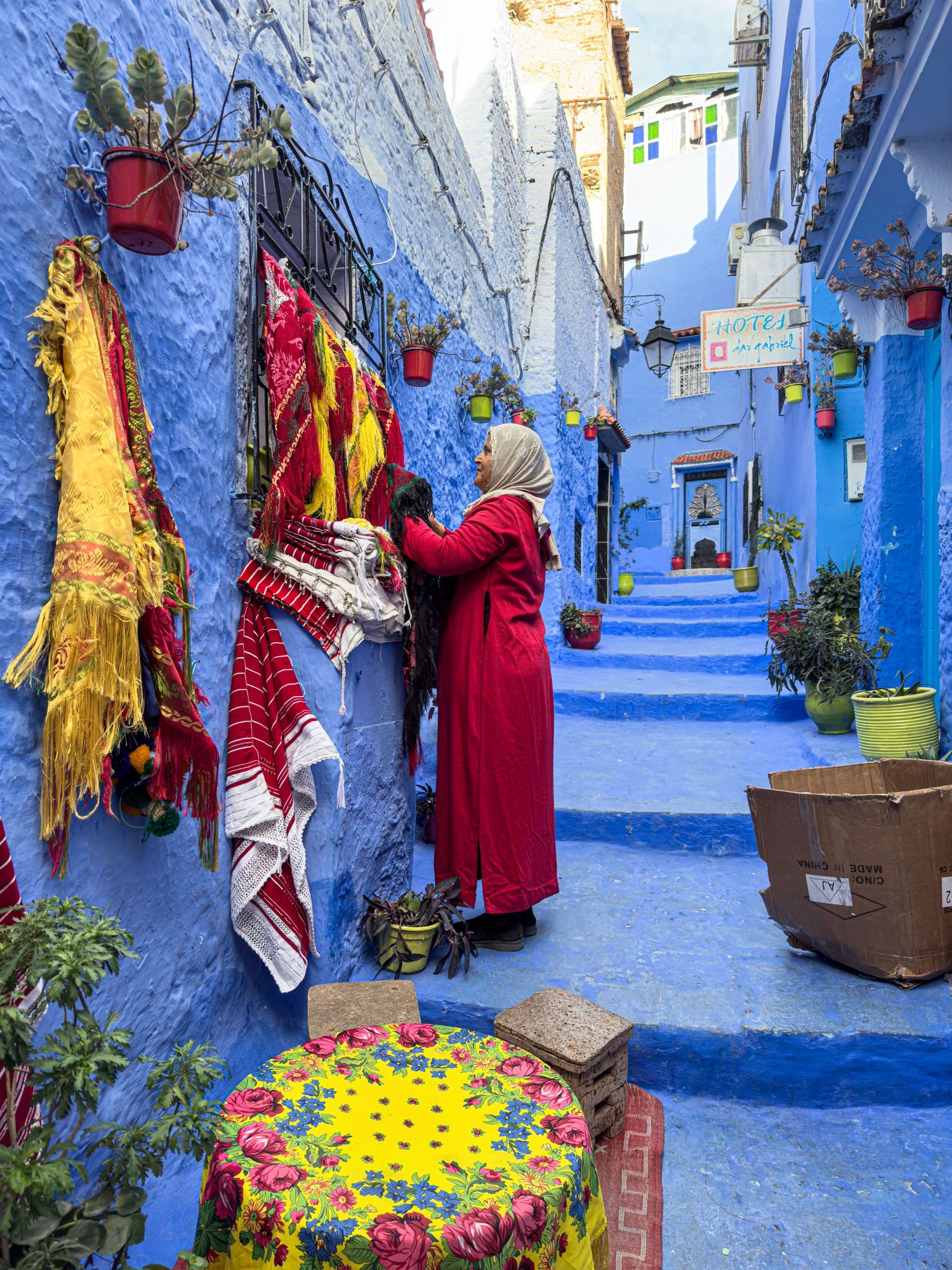 Woman in red coat browsing colorful textiles in a vibrant blue alley of Chefchaouen, Morocco.