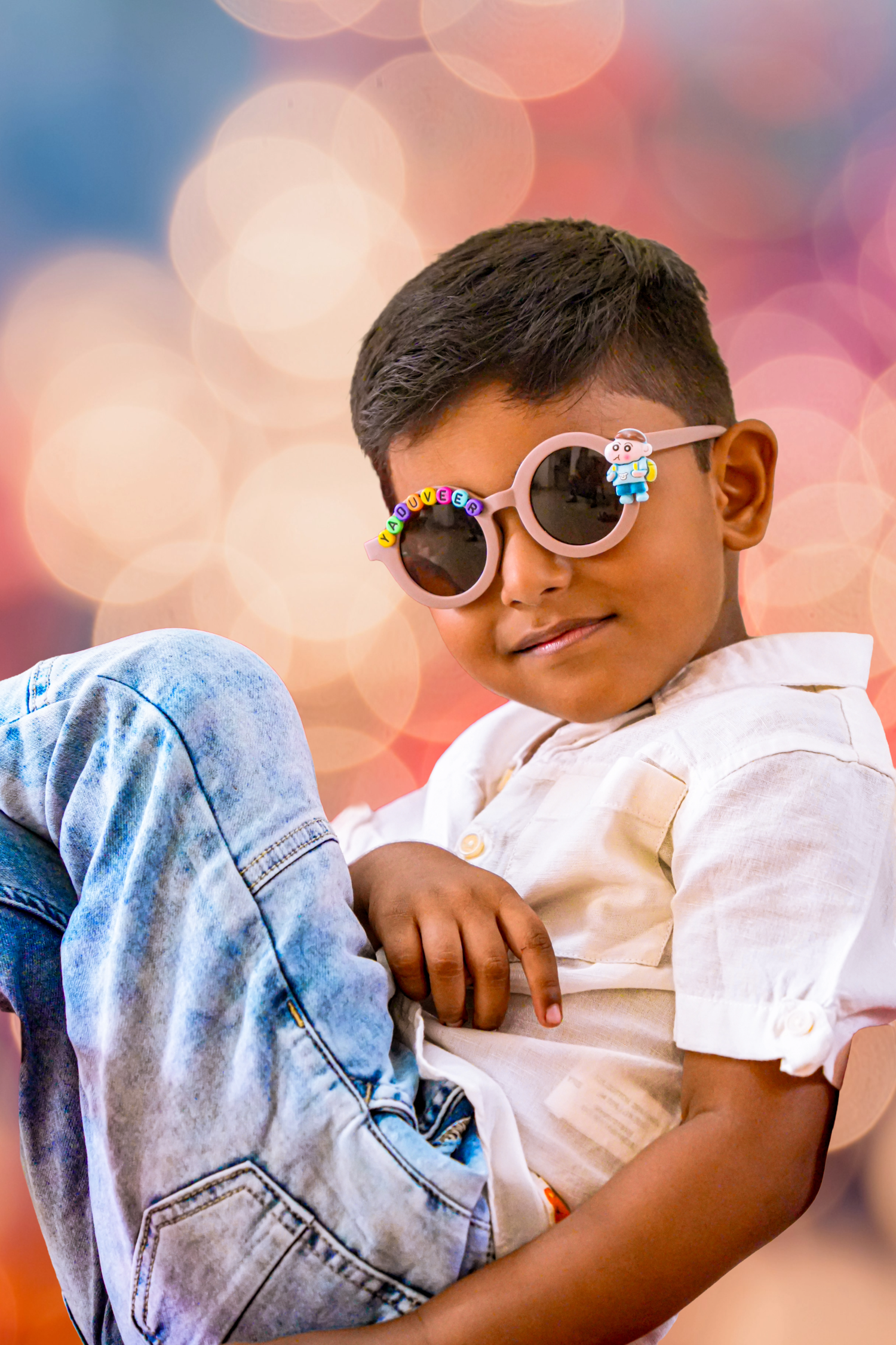 Cute Indian boy wearing decorative round sunglasses and white shirt sitting casually with bokeh background.