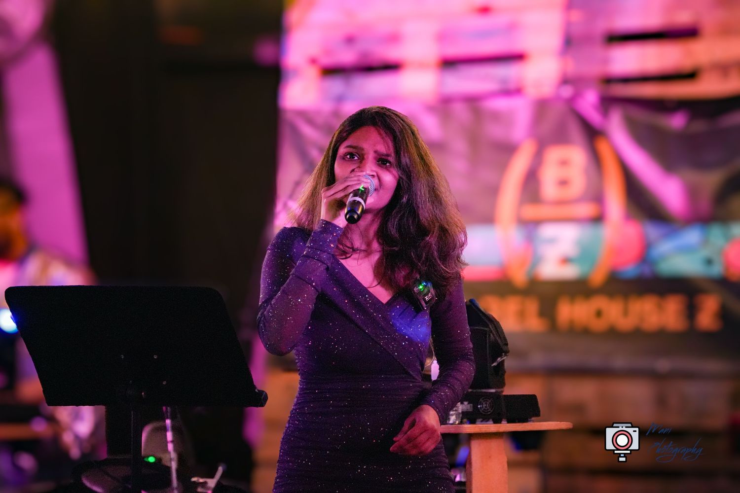Woman in sparkly blue dress singing into microphone on stage under pink lights at a live music event.