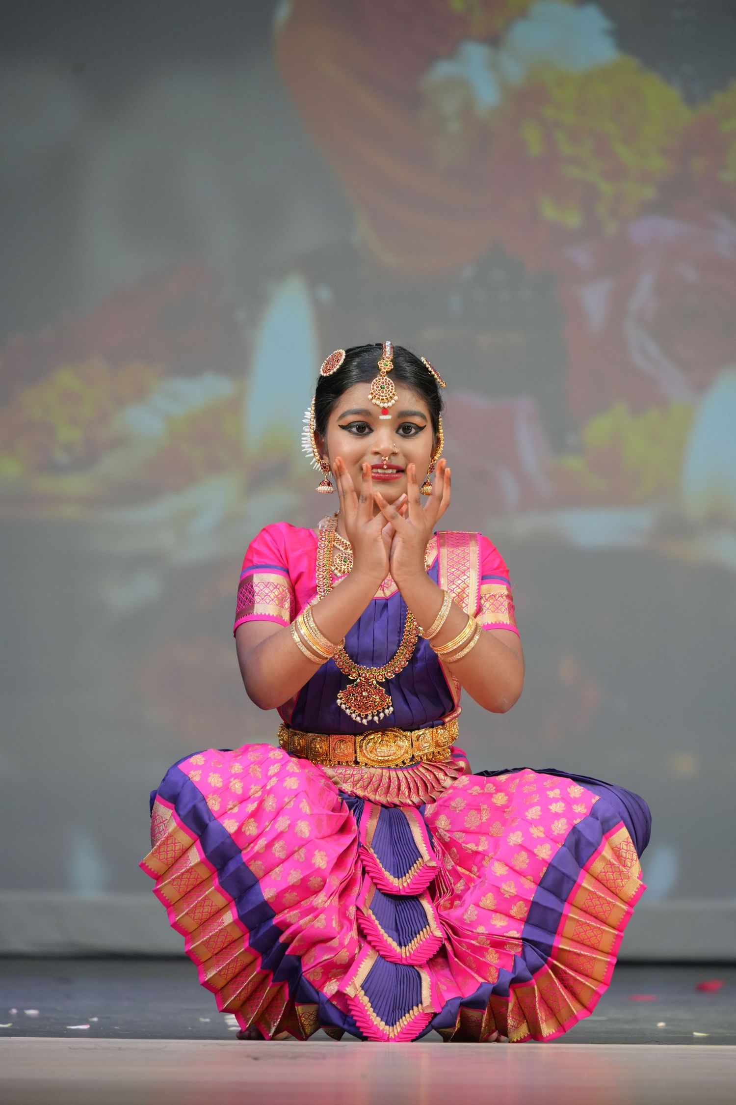 Young girl performs Bharatanatyam classical Indian dance in vibrant pink and purple silk costume with gold jewelry on stage.
