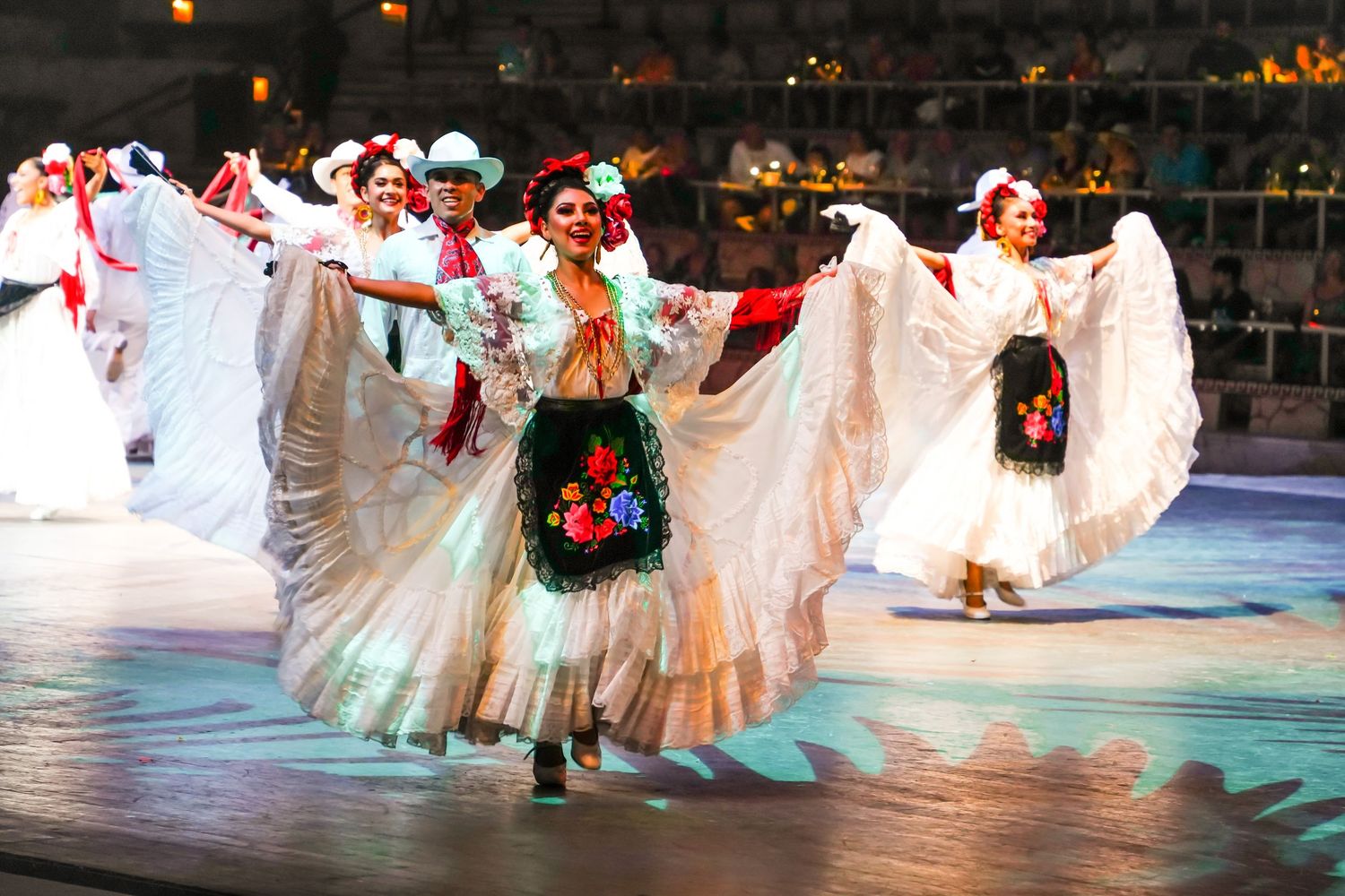 Colorful Mexican folk dancers in white floral dresses perform traditional Folklórico dance on a lit stage.