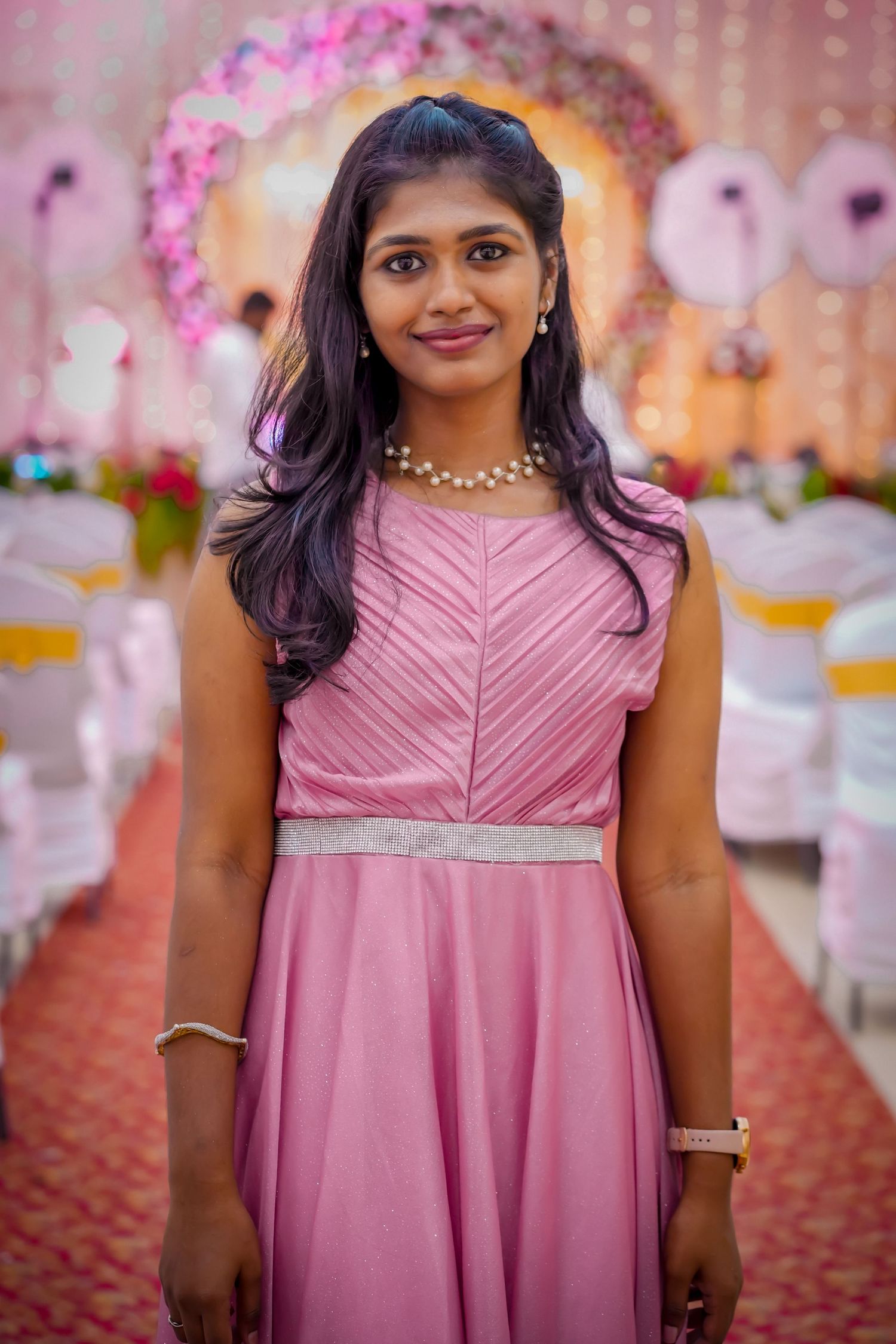 Young Indian woman in a pink gown with pearl necklace smiling at a beautifully decorated wedding venue.