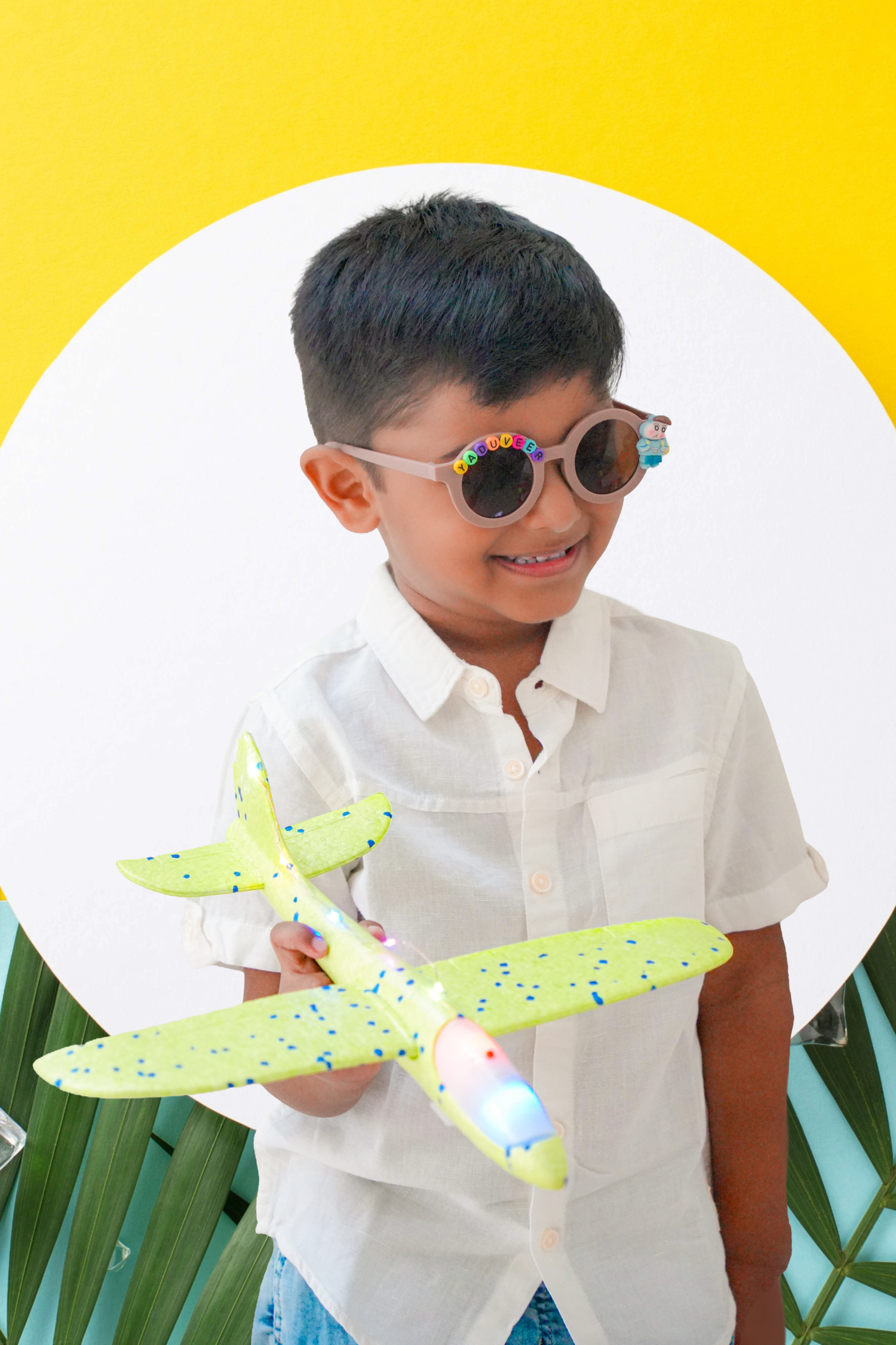 Happy Indian boy in white shirt and round sunglasses holding a green toy airplane.