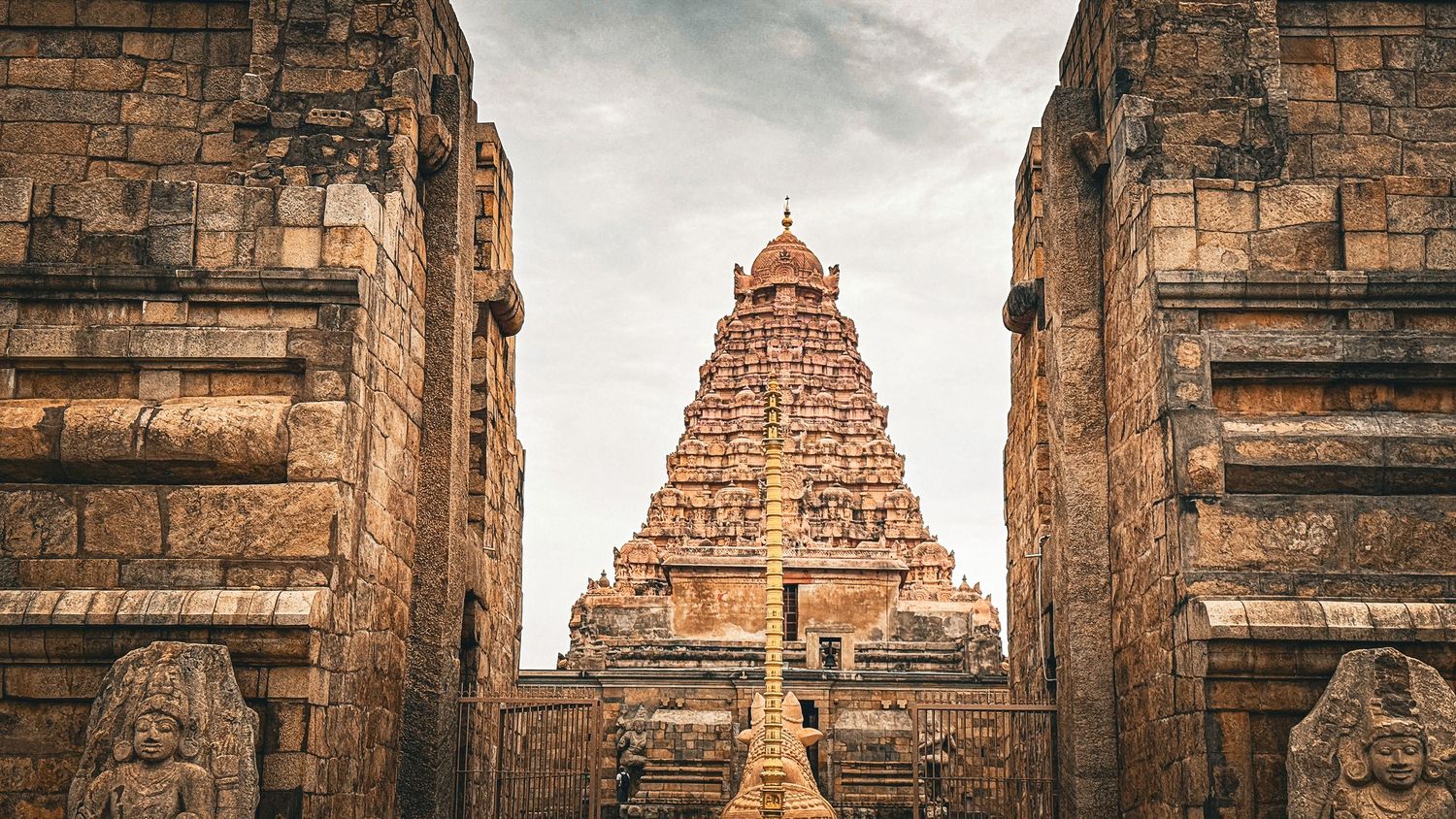 Ancient Brihadeeswarar Temple gopuram framed by stone pillars under dramatic cloudy sky in Thanjavur, India.